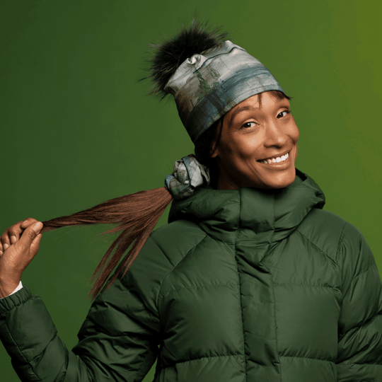 A smiling woman wearing a green winter coat and the Wall Beanie With Pompom pulls her ponytail.