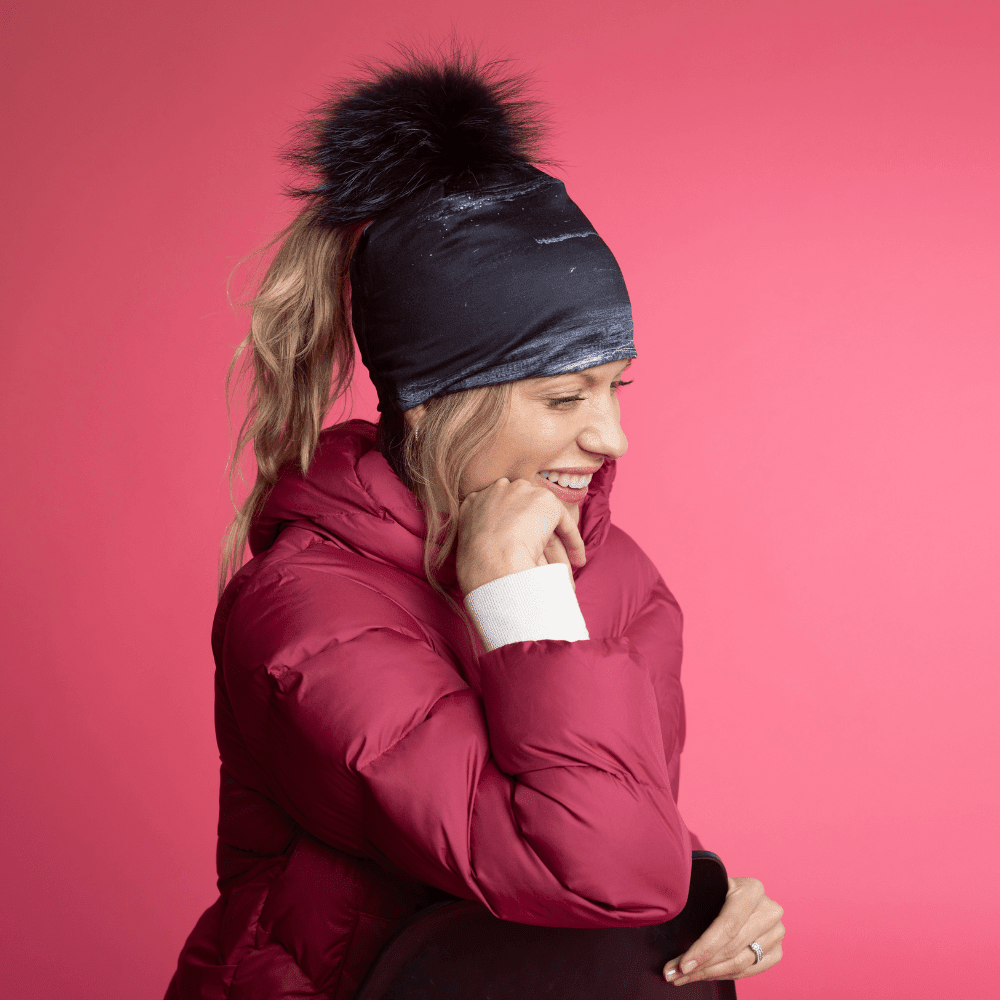 Woman in a red puffer jacket and the Wall Black Ponytail Beanie, smiling and looking down against a pink background.