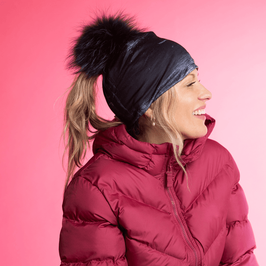 Side profile of a woman wearing the Wall Black Ponytail Beanie and a red puffer jacket against a pink background.