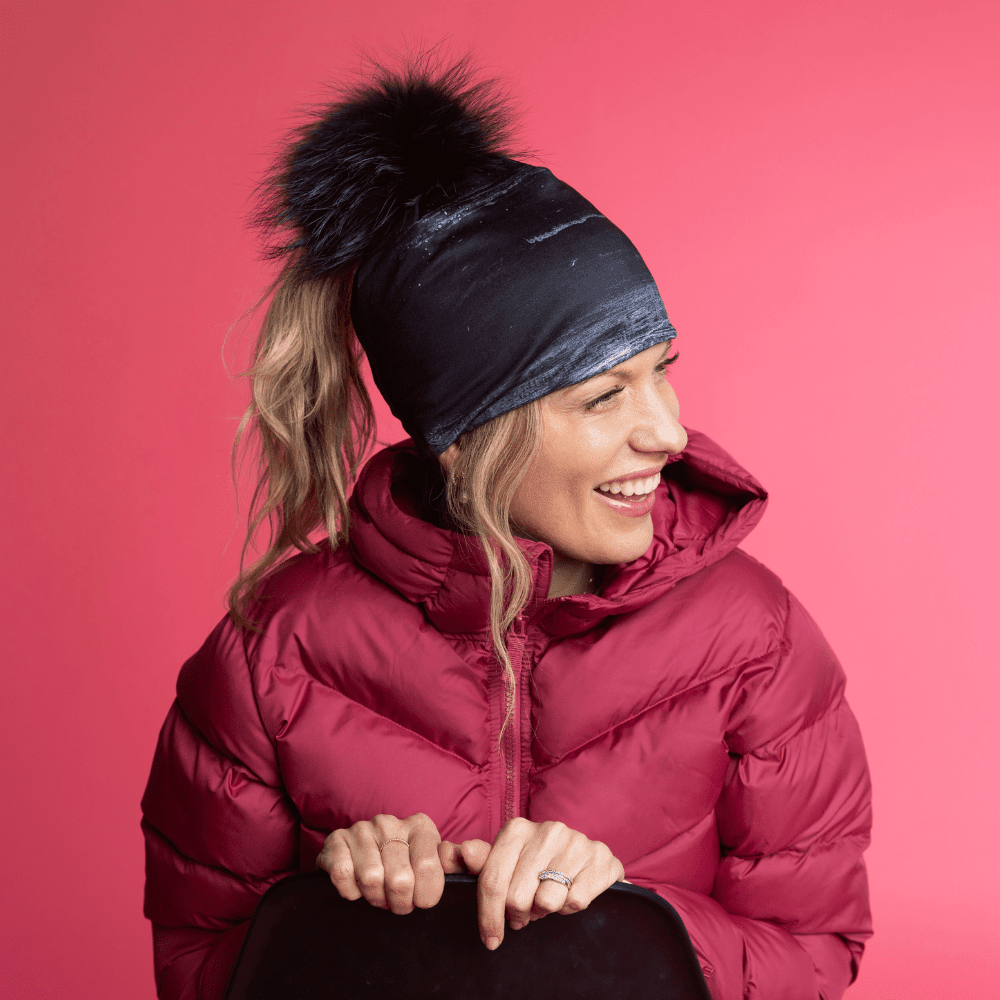 Smiling woman wearing the Wall Black Ponytail Beanie and a red puffer jacket against a pink background.