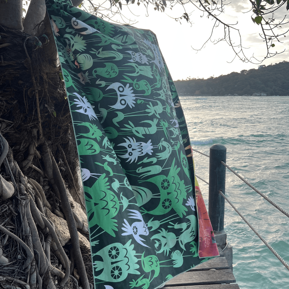 The Wall of Birds towel hangs from a tree branch near the ocean, displaying its dark green side with white and light green bird illustrations.