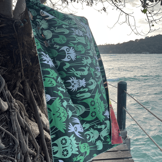 The Wall of Birds towel hangs from a tree branch near the ocean, displaying its dark green side with white and light green bird illustrations.