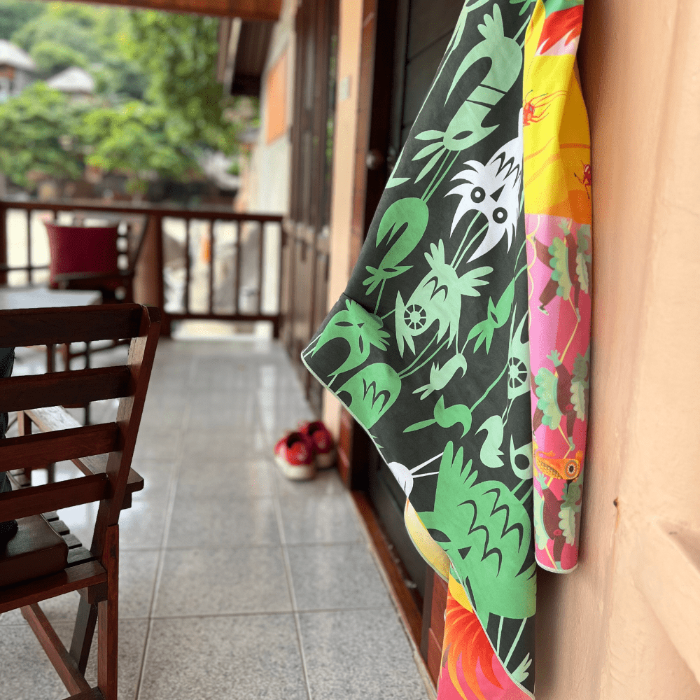 The Wall of Birds towel, showing both colorful and dark green sides, is casually hung on a textured wall outdoors.