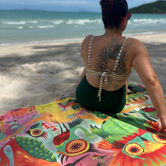 A woman sits on a sandy beach looking out at the ocean, with the colorful side of the Wall of Birds towel spread out before her.