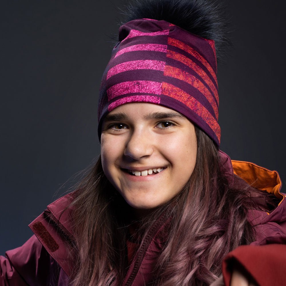 Close-up of a smiling young woman wearing a purple and pink striped beanie with a black pom-pom.