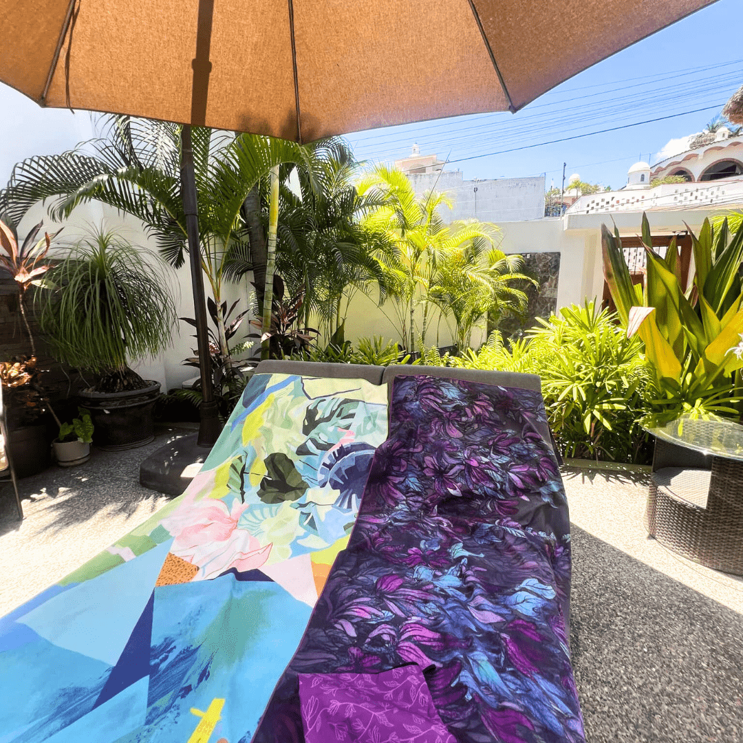 Wisteria Luxury Microfiber Towel and another art towel draped on outdoor lounge chairs under a sun umbrella.