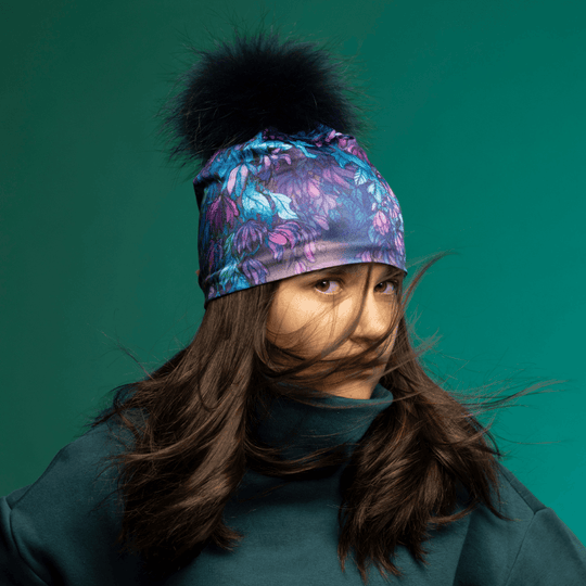 Young woman wearing the Wisteria Pompom Beanie, with her hair blowing in the wind against a teal background.