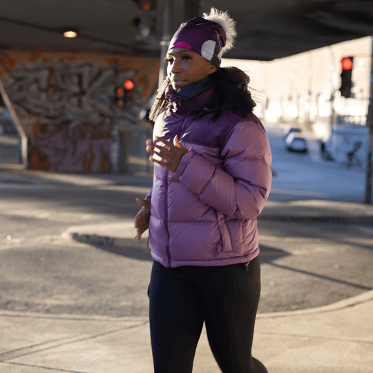Side profile of a woman running outdoors, wearing a magenta beanie with a gray pompom and a grey neck warmer.