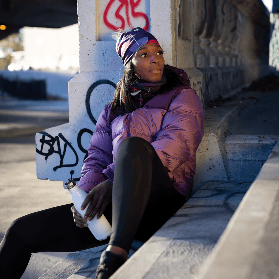 Woman sitting outdoors under a concrete structure, wearing a purple puffer jacket and a magenta beanie with a gray pompom, holding a white water bottle.