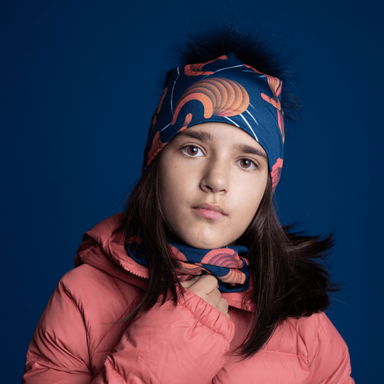 Close-up of a young girl wearing the navy beanie with orange pattern and a matching neck gaiter, adjusting the gaiter