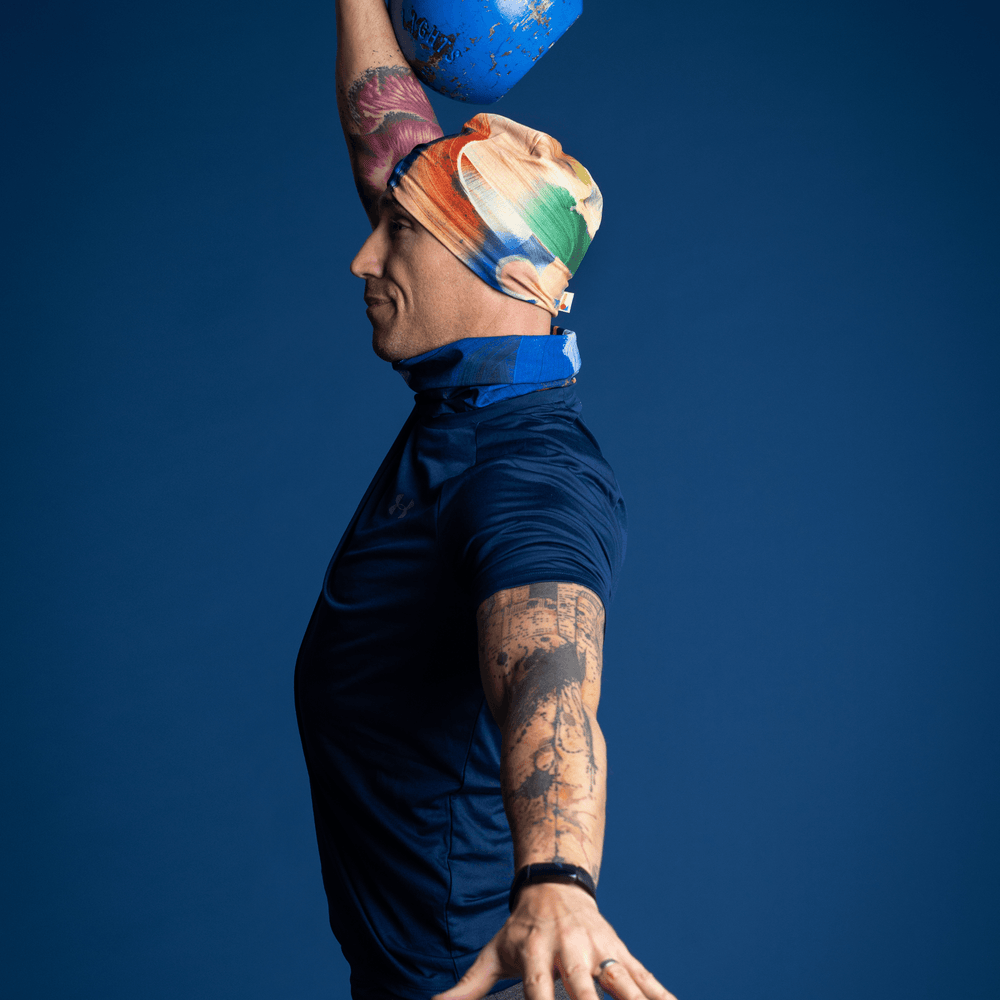 Man wearing the yellow and blue abstract art beanie and matching neck tube, performing a kettlebell clean.
