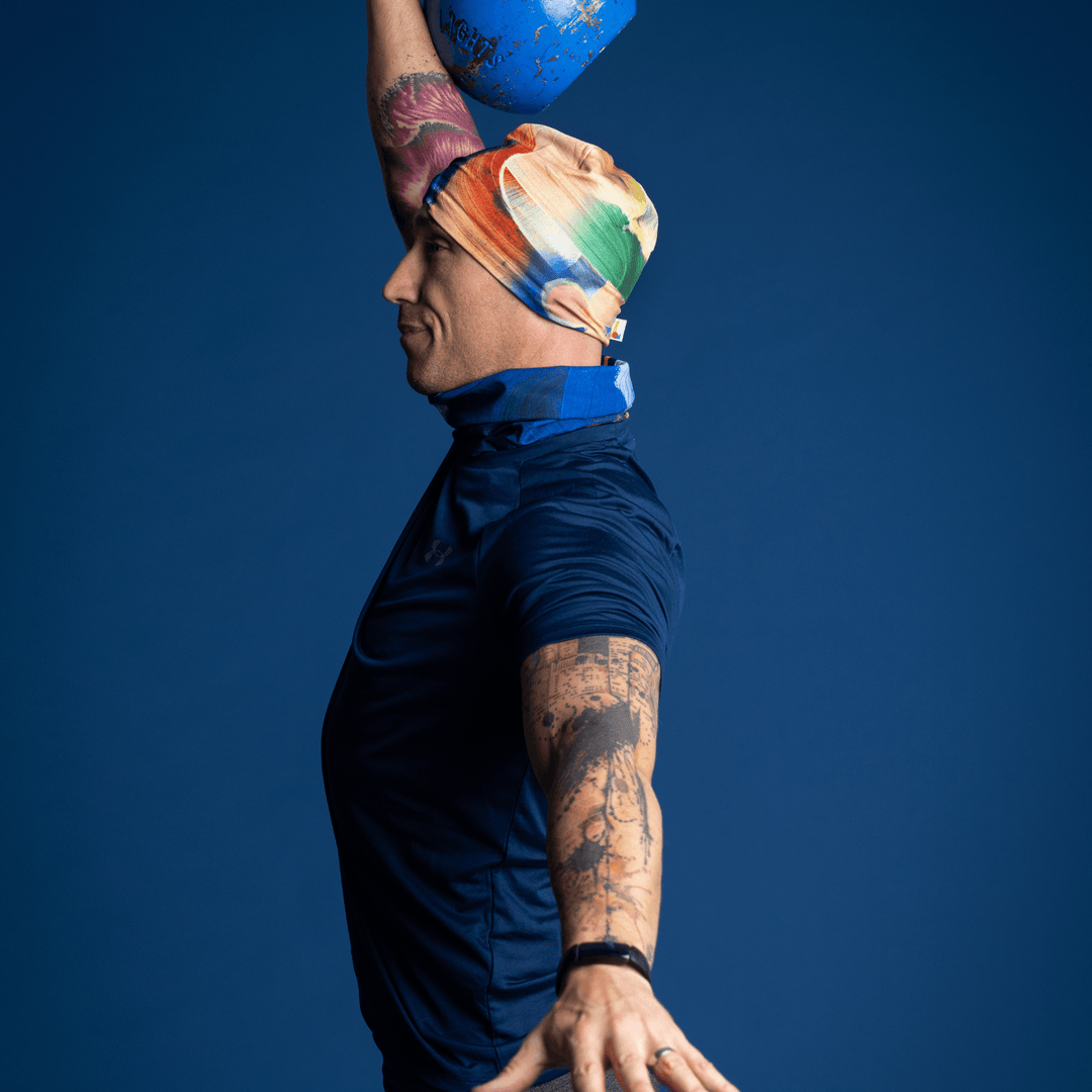 Man wearing the yellow and blue abstract art beanie and matching neck tube, performing a kettlebell clean.
