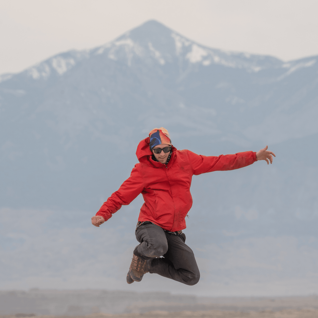 Person in a red jacket leaping in the air against a mountainous backdrop, wearing the abstract art beanie.