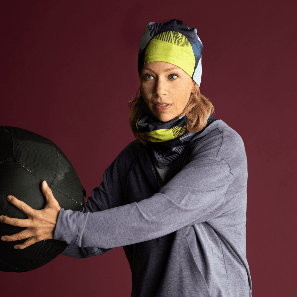 Woman wearing a gray long-sleeved shirt and the Yellow Dots neck tube as a headband and around her neck, holding a large black ball