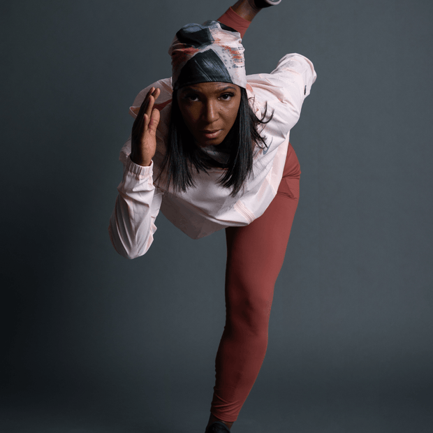 Woman in front running and is in motion. The woman is wearing a 4 seasons yoga hat designed by Catherine Parent. The hat is colored in pink, grey and beige tones, and has a black pompom. 